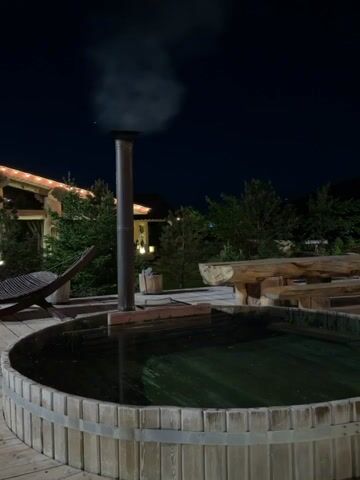 The video captures an outdoor nighttime scene featuring a wooden hot tub or plunge pool. In the foreground, a circular wooden hot tub is visible, filled with dark water. A tall, silver metal chimney pipe rises from the center of the tub, emitting a steady stream of white smoke, indicating that the tub is being heated. The hot tub is surrounded by a wooden deck.

To the left of the hot tub, on the wooden deck, there is a dark, slatted wooden lounge chair. In the background, lush green trees and bushes are scattered, suggesting a natural or garden setting. Behind the foliage, a building with warm string lights adorning its roofline and windows is illuminated, creating a cozy ambiance against the dark night sky. Further to the right in the background, a rustic wooden table and benches, made from large logs, are visible. The overall scene is dark, characteristic of nighttime, with artificial lights providing illumination to the structures and some areas of the ground. No people are present in the video.