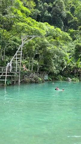 This media file captures a vibrant scene at a tropical swimming hole, likely in Ban Nampoun, Laos. The water is a striking turquoise, surrounded by lush, dense green foliage that cascades down from a steep hillside. In the background, banana trees and other tropical vegetation thrive under what appears to be a bright, sunny day, suggesting a warm, humid climate.

The primary activity is recreational swimming and adventure. On the left, a wooden platform with a ladder stands tall, leading to a zipline. A person is shown mid-zipline, gliding over the water. In the water, several individuals are enjoying the cool, clear water. Two people are floating on inner tubes, relaxed and chatting, while another person swims nearby. The overall mood is one of leisure and enjoyment in a natural, beautiful setting.

A notable detail is a weathered wooden sign partially obscured by foliage, with the text "BLUE MOON" clearly visible. This likely indicates the name of the location or a specific attraction within it. The clear water and vibrant greenery create a picturesque and inviting atmosphere, highlighting a popular spot for outdoor recreation and relaxation. The scene is bright and suggests it is midday or early afternoon due to the quality of light.