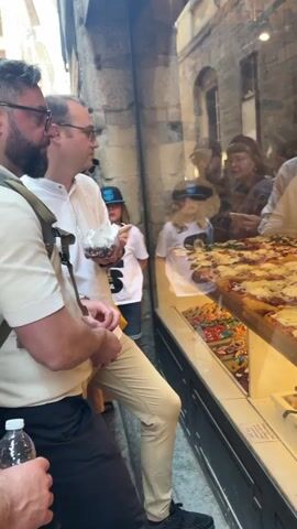 People are looking at pizzas on display in a glass case, likely outside of a restaurant or a bakery. Some of the people in the shot are reflected in the glass. The pizzas are rectangular and have different toppings, including mozzarella, ham, vegetables, and hot dogs. A woman wearing an apron and black gloves is behind the case cutting a slice of pizza. She appears to be working in the restaurant. The environment looks like a narrow street with stone buildings. There's also a small price tag visible in the bottom right corner of the glass case.