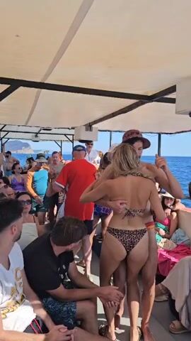 The video shows a crowded boat filled with people enjoying a sunny day on the water. The camera focuses on a man and woman in the center, who are embracing and appear to be dancing. The woman is wearing a leopard print bikini, and the man is wearing swim trunks and a hat. Other passengers are visible in the background, some sitting and some standing, all dressed in casual, summer attire. The boat has a canopy providing shade, and the blue ocean and distant coastline are visible. The atmosphere seems lively and festive.