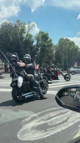 The video shows a group of motorcyclists stopped at a city intersection. There are several other vehicles visible on the road, including cars and a red motorcycle. The motorcyclists are waiting for the traffic light to change. The first motorcycle in the group has a flag that says "Floyd" on it. The second motorcycle in the group has a sidecar. The third motorcycle in the group is a scooter. In the background, there are tall buildings and trees. The sky is blue and sunny. The video is shot from inside a car, looking out through the windshield.
