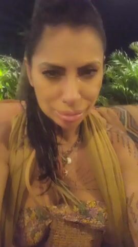 A woman is seated outside at night, drinking from a clear glass with a straw. She has dark hair pulled back, fair skin, and visible tattoos on her chest and arms. She is wearing a floral top and a loose, yellow wrap. The drink in the glass is orange. Lush green foliage is visible in the background. The woman appears to be talking to the camera while she drinks.