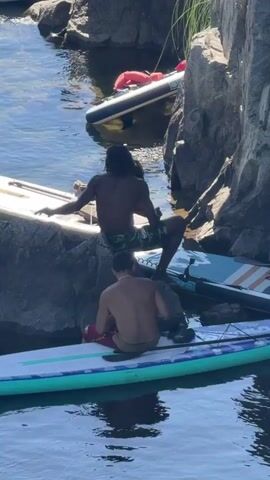Two individuals are on paddleboards near rocky outcrops in a body of water. One individual, with dark hair and wearing patterned shorts, is sitting on a rock with their back to the camera and appears to be kissing another individual who is partially obscured. Below them, another individual wearing red shorts is sitting on a paddleboard. Several other paddleboards are visible around the area. The water is blue and reflects the sunlight. Other people are visible sitting on rocks and paddleboards in the background. The overall scene suggests a recreational outing on the water.