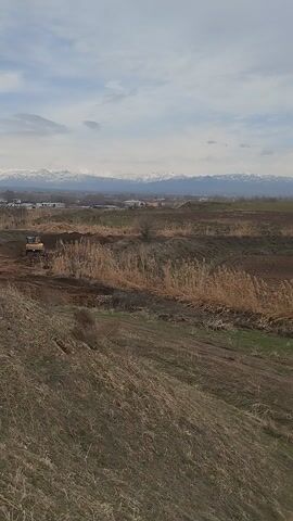 The video opens with a wide shot of an open, somewhat barren landscape. In the mid-ground, a yellow excavator is visible, actively engaged in earthwork. It moves forward, its shovel seemingly digging into or pushing soil, creating or deepening a trench-like depression in the brown, dry ground. The immediate foreground is covered in dry, light brown grass and patches of bare earth, indicating a fallow field or undeveloped land.

In the background, a majestic range of mountains stretches across the horizon, their peaks dusted with snow, suggesting a cooler climate or higher elevation. The sky above is overcast with white and light gray clouds, diffusing the daylight. In the distance, between the field and the mountains, hints of human settlement—possibly buildings or structures—can be vaguely discerned, suggesting the area is not entirely remote.

As the video progresses, the excavator continues its work for a few seconds before moving further to the right and eventually out of the frame. The camera remains relatively still, focusing on the landscape. The latter part of the video shows only the dry, uneven field, with some faint green patches emerging, and the distant, snow-capped mountains under the cloudy sky. The overall impression is one of ongoing development or agricultural preparation in a scenic, mountainous region.