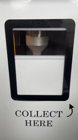 This is a close-up, vertical video showing a soft-serve ice cream machine dispensing ice cream into a paper cup. The machine is white with a black frame around the dispenser window. Below the window, the text "COLLECT HERE" is printed in black capital letters, with an arrow pointing to the right.

The video begins with the empty dispenser nozzle visible above the empty paper cup. In the subsequent frames, soft-serve ice cream is dispensed into the cup, topped with colorful candy pieces. The ice cream appears to be a light yellow or vanilla flavor. The setting is indoors, likely a shop or kiosk, given the context of an ice cream machine. There are no people visible in the video. The lighting appears to be artificial and consistent throughout, suggesting it could be any time of day. No specific weather or emotional cues can be determined from the video.