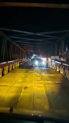 The video shows a nighttime view from inside a vehicle, looking out through the windshield at a bridge. The bridge appears to be made of metal trusses and has a wooden or metal deck. Bright yellow lights illuminate the bridge deck and a yellow car with its headlights on is stopped in the middle of the bridge. A person in a yellow vest is standing next to the car. Further down the bridge, other vehicles and lights are visible, suggesting a traffic situation. The overall environment is dark, indicating nighttime. The car's license plate is partially visible.