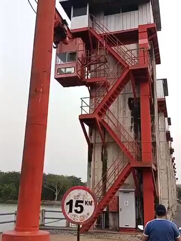 The video shows a large red industrial structure with a crane and a control booth at the top. A red staircase leads up the side of the structure. Below the structure, there is a body of water and a fence. A circular speed limit sign showing "15 KM" is visible in the foreground. A person is standing with their back to the camera, looking towards the water. The sky is overcast.