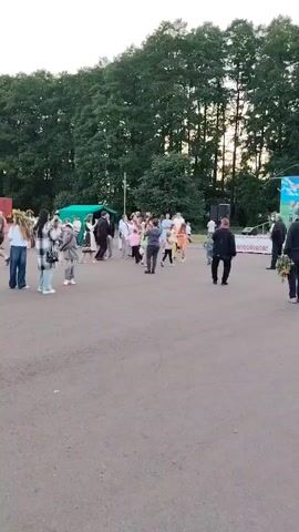 The video shows an outdoor gathering with a group of people dancing. The setting appears to be a paved area, possibly a community space, with a line of trees in the background. The lighting suggests it is late afternoon or early evening, with the sun casting warm light through the trees. Some individuals are dressed in what appear to be traditional or festive clothing, indicating a cultural event or celebration. Children are actively participating in the dancing, and adults are also engaged, some observing while others join in. There are speakers set up, suggesting music is playing, contributing to the lively atmosphere. The overall mood seems joyful and communal. No visible text is present in the video. The location context provided is Олтушскі сельскі Савет, Belarus, which helps place the event within a specific rural Belarusian community.