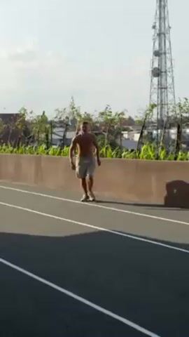 A shirtless man wearing khaki shorts is jogging on a black track with white lines, next to a low concrete wall with greenery, and a tall metal tower in the background. He performs a breakdance move, spinning on his hands and feet. The man stands up, looks at the camera and smiles. A strip of athletic tape covers the bridge of his nose. He pivots away from the camera, revealing a mullet hairstyle.