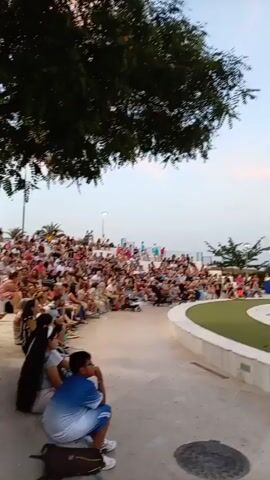 This media file captures an outdoor performance or rehearsal, likely a dance recital, taking place in an amphitheater or similar tiered seating area. The scene is set at dusk, with the sky displaying soft, pastel hues of pink and blue, indicating either sunrise or sunset. The overall mood appears serene and anticipatory, with the audience seated on the tiered levels, facing a central performance space.

The performance area is a large, open square with a checkered floor pattern of light and dark squares, resembling a giant chessboard. A few performers are visible in the center of this space, dressed in light-colored, flowing attire, suggesting a dance or dramatic piece. Some are in dynamic poses, while others appear to be resting or preparing for their next move. In the background, a crowd of spectators fills the tiered seating, their faces indistinct due to the distance and lighting.

Several large, black speakers are strategically placed on the performance floor, indicating that music or amplified sound is integral to the event. The setting is semi-urban, with buildings and houses visible on a hillside in the background, a common sight in many Spanish towns. The presence of trees framing the top left of the frame adds a natural element to the urban landscape. The ground in the foreground is paved, with a circular metallic grate visible, suggesting a public space. The weather appears clear and pleasant, conducive to an outdoor gathering.

While no explicit text is visible in the media, the context provided, "Olula del Río, Spain," places the event in a specific geographical location. The image evokes a sense of community and artistic expression, common in local cultural events.