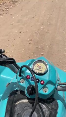 This media file opens with a close-up view of a turquoise ATV's dashboard. The odometer shows a speed of approximately 10 km/h, with the engine in neutral ("N"). A "Park Off" indicator is illuminated. The scene then shifts to a wide shot of a dry, grassy field under a hazy, overcast sky. In the distance, a barren, hilly landscape is visible. The camera then focuses on a camel with a red rope around its neck, standing in the field. The camel appears to be in a rural, possibly desert-like environment. The overall impression is one of outdoor exploration or travel in a remote, arid region. The text visible on the ATV dashboard includes symbols for a light bulb, a snowflake, a headlight, and indicators for neutral ("N") and reverse ("R"). A yellow tag is visible on the camel's ear.