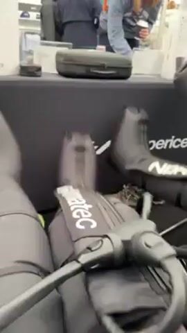 The video shows a close-up of Normatec compression boots inflating and deflating on a table at what appears to be a public event. The black boots are labeled "Normatec" and "Normatec C". In the background, several people browse a table with various items displayed. A woman in a baseball cap is holding a coffee cup and examining a small object in her hand. Another person wearing a jacket is standing beside her. The table is covered in black fabric with the word "Experience" printed on it. The scene is brightly lit, suggesting an indoor setting.