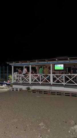 The video shows a beachfront cafe at night. People are seated on a deck, watching a television screen that displays a soccer match. The cafe is illuminated by lights, and the surrounding area is dark. The ground in front of the cafe is sandy.