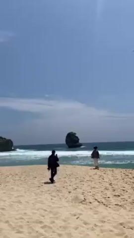 The video captures a beach scene on a sunny day. The sky is clear with a few scattered clouds. The sand is light-colored. The ocean has waves breaking near the shore. There is a large rock formation topped with greenery visible in the ocean. Two people are standing near the shoreline. One is walking away from the camera. Another person is walking into the frame from the left and towards the right. A pink cap is also visible entering the frame.