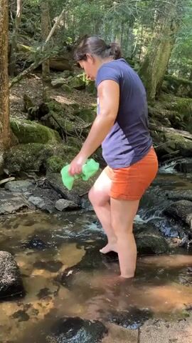 A woman with dark hair pulled back stands in a shallow stream. She is wearing a dark blue t-shirt and orange shorts. She is holding a green cloth in her right hand and is moving it through the water. Her bare feet are visible in the water, which is clear and flowing over rocks. The surrounding environment is a forest with trees, moss-covered rocks, and fallen logs. Sunlight filters through the trees, creating dappled light. The woman appears to be rinsing the cloth in the stream.