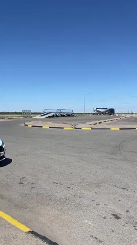 The video depicts a paved area with a section marked off by black and yellow road markers. In the distance, a semi-truck is parked. The camera pans to reveal a white car carrier truck parked to the right of the frame, with its driver's side door open. The sky is a clear, bright blue. There are streetlights lining the edge of the paved area. Other vehicles can be seen driving on a highway in the distance.