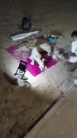 A group of people are gathered on a sandy surface, possibly a beach or desert, illuminated by a bright light. In the center, a pink cloth serves as a base for several clay or plaster sculptures of animals, appearing to be unfinished. A person in a white shirt and brown pants is seated, actively working on one of the sculptures. Next to them, a red and white pole extends upwards, possibly part of a camera or lighting setup. Scattered around the area are various objects including a black bag, white fabric, and other unidentifiable items. The ground is uneven and sandy, with some debris visible. The scene suggests an outdoor artistic endeavor taking place in the evening or night.