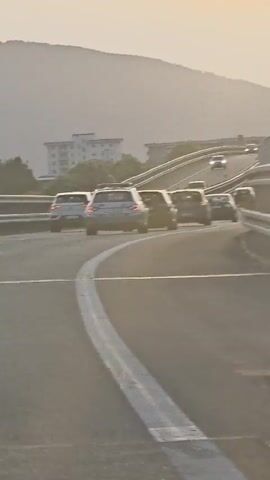 A convoy of cars is traveling on a highway that curves to the left. The cars are moving away from the camera on an upward incline. The sky is hazy and the sun is low in the sky, suggesting it is either sunrise or sunset. The sun is a bright, diffused circle in the upper part of the frame, casting a warm glow over the scene. The highway is lined with guardrails. In the background, rolling hills can be seen.