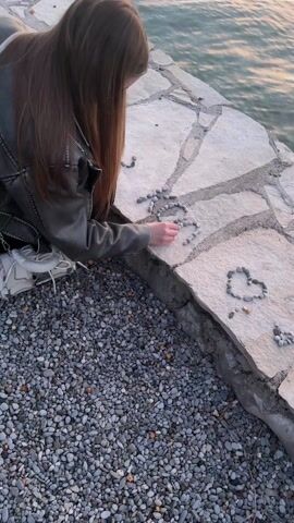 Here's a description of the video:

A young woman with long, reddish-brown hair is kneeling by the water's edge, arranging small pebbles on a stone surface. She is wearing a gray jacket and white sneakers. She appears to be creating shapes, including a heart, with the pebbles. The stone surface is part of a walkway or wall next to a body of water. There are more pebbles on the ground around her. In the background, the water reflects the colors of the sunset, which is visible on the horizon along with a line of hills.