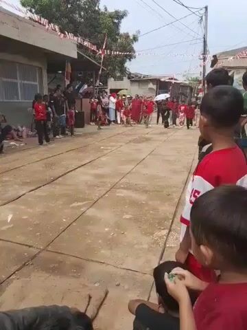 The video shows an outdoor street celebration. In the foreground, several children, mostly wearing red and white, are watching an event. In the background, a race is taking place on a dirt path lined with square paving stones. Participants are running, and one person is carrying an object, possibly a sack. Spectators line the sides of the path, some seated and others standing. Red and white Indonesian flags are strung across the street, decorating the buildings. The atmosphere appears festive.