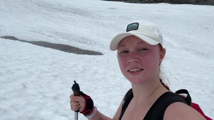 A young woman wearing a white cap and a black tank top is filming a selfie video outdoors in a snowy, mountainous landscape. She has reddish-brown hair in a braid and is smiling at the camera. She holds a trekking pole in her right hand. In the background, a group of people are gathered on a rocky, snow-covered slope. The sky is overcast.