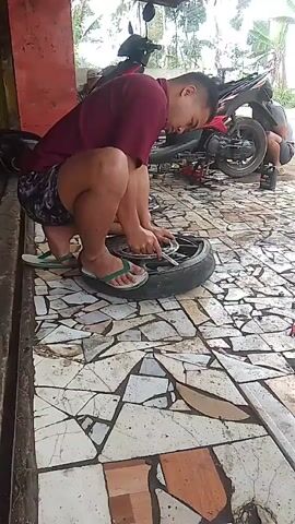 A man in a maroon short-sleeved shirt and patterned shorts is kneeling on a tiled floor, working on a motorcycle tire. He is using a tool to remove something from the wheel hub. His left foot is bare, and he is wearing a green flip-flop on his right foot. Behind him, a red motorcycle is partially visible, propped up on a stand, with another person's legs visible underneath it, also working on the motorcycle. The environment appears to be an outdoor workshop or garage, with a roof overhead and trees visible in the background. The tiled floor is broken in places, with a mosaic-like pattern.