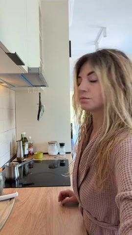A woman with blonde, wavy hair, wearing a pink bathrobe, stands in a kitchen. She is facing away from the camera, towards a black stovetop. On the stovetop, a small metal pot is visible. The woman is holding a spoon in her right hand and appears to be interacting with the pot or the stovetop. The kitchen counter is made of wood, and there are various bottles and containers on it, including what appears to be olive oil and a jar of salt. A stainless steel range hood is mounted above the stovetop. The background shows a doorway leading to another room, with light streaming in from a window.