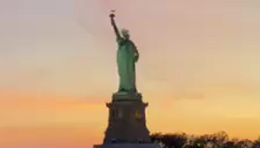 The media file displays a full-body shot of the Statue of Liberty, a monumental sculpture, silhouetted against a sunset sky. The sky transitions from a pale pink near the horizon to a soft grey at the top, with hints of orange and yellow in between, suggesting it is either sunrise or sunset. The Statue of Liberty stands tall on its pedestal, its torch held aloft. The sculpture appears to be made of copper, exhibiting a greenish patina. In the background, the dark shapes of trees are visible, adding depth to the scene. The lighting indicates that the photograph was taken during the twilight hours. No people are discernible in the frame, and there is no visible text. The setting is clearly Liberty Island in New York Harbor, given the iconic subject. The overall mood conveyed is serene and majestic, characteristic of a sunset view of this famous landmark.