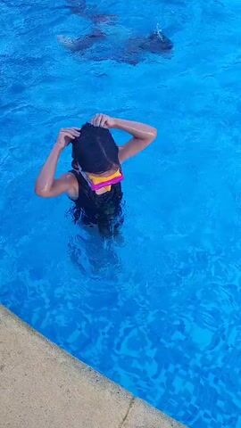 The video shows two children in a blue swimming pool. The child in the foreground is wearing a black swimsuit and yellow and pink goggles. They are standing in the water up to their chest and have their hands raised. Another child is partially submerged in the background, with their legs visible above the water. The water is clear and blue, with ripples on the surface. The edge of the pool, a concrete-like material, is visible in the lower left corner of the frame. The camera is positioned above and to the side of the children, looking down into the pool.