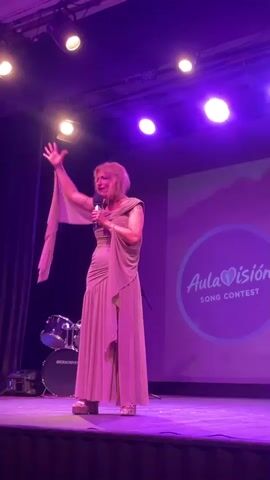 The video shows a woman on stage in a light pink dress, holding a microphone and gesturing with her left hand. She is standing in front of a large screen that displays "Aula Visión Song Contest" with a heart symbol. To the left of the stage, there is a drum set and a stool. The stage is lit with purple lights. The woman appears to be performing or presenting.