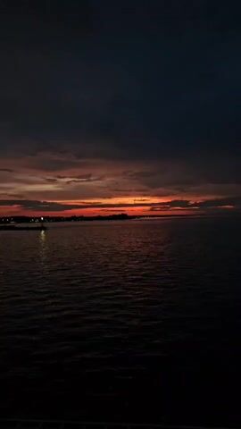 The video shows a body of water at dusk. The sky is dark with some clouds, and there is a band of orange and red near the horizon, suggesting a sunset. Lights from a distant shore are visible on the left side of the frame, reflecting on the water. The water is dark and rippling.