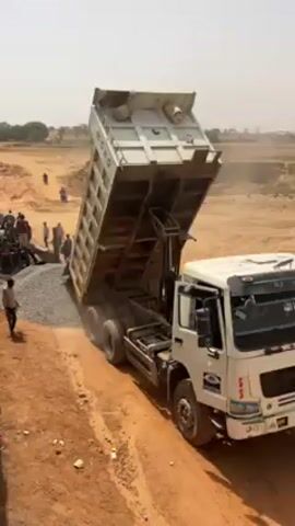 Here is a description of the video.

The video shows construction work, most likely on a bridge or overpass. A dump truck unloads gravel onto a dirt road. People stand around, possibly overseeing or assisting. A man walks by. The camera pans to show workers spreading fresh concrete over a metal rebar grid on a raised platform. In the background is a rural landscape with trees and fields.