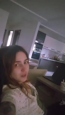 A woman is lying on a couch, facing the camera. She is wearing a white shirt with a green pattern. Her hair is dark and shoulder-length. She puts her right arm up and behind her head. There is a dark couch behind her and a messy side table on the right. In the background, there is a dining room table, a kitchen, and a wall decorated with mirrors and framed artwork. Music is playing.