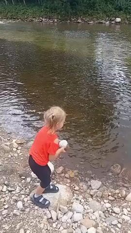 The video captures a young girl with blonde hair pulled back in a ponytail, wearing a bright orange t-shirt with cut-out shoulders and black leggings. She stands on a rocky shoreline next to a river. The girl is in the process of throwing a light-colored rock into the water, which creates a splash. The river is calm with visible ripples and the opposite bank is lined with trees and greenery. The shoreline is covered in various sizes of grey and white stones.