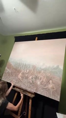 A woman with long, curly hair, wearing a dark top, is seen adjusting a very large painting on a wooden easel. The painting, which occupies most of the frame, is rectangular and depicts a somber, desaturated scene.

The artwork features a group of figures, possibly human, gathered in the foreground amidst a desolate, rugged landscape. The color palette is dominated by earthy browns, grays, and muted blues, with a pale, possibly pinkish-beige sky. Some figures are seated, one appears to be holding a staff, and another holds what looks like a glowing object near the ground. The background is filled with what seems to be ruins, debris, or a chaotic, heavily textured environment, suggesting a post-apocalyptic or ancient setting. The overall mood of the painting appears serious or contemplative.

The woman is positioned to the left of the canvas, pushing it upwards on the easel. At the beginning of the video, there's a distinct clattering sound as the heavy canvas momentarily slips or adjusts, but she quickly regains control and continues to reposition it. The setting appears to be an indoor room, with a green wall to the left and a black curtain behind the easel. The ceiling is white. There is no visible text in the video.