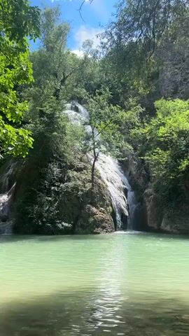 A serene video captures a natural waterfall cascading down a rocky cliff face into a clear, turquoise pool of water. Lush green foliage surrounds the waterfall and frames the shot, with sunlight filtering through the trees, creating dappled patterns on the water's surface. The water flows gently from the pool, disappearing into the lower foreground of the frame, hinting at a wider river or stream beyond. The scene evokes a sense of peace and natural beauty, with no people or man-made objects visible.