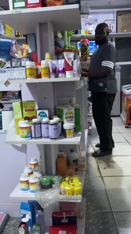 The video depicts a man standing in a small store, possibly a pharmacy or general store, filled with shelves displaying various products. The man has dark skin, a beard, and is wearing a grey short-sleeved shirt with white stripes on the sleeves and black pants. He's standing near a white shelving unit stocked with items such as medications, supplements, and personal care products. Some visible brands include "Calcitone," "Nerve," and "GOKO." The shelving unit is white and appears to be made of particle board. The floor is covered in light-colored tiles. Behind the man, there is a small white freezer and an orange plastic stool. The store has a computer monitor and other assorted items on higher shelves. The lighting in the store is bright and somewhat artificial. The camera moves slightly during the recording, providing different views of the store interior.