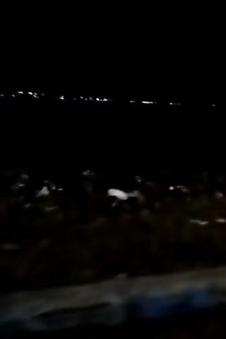 The video is a low-resolution, shaky night-time recording. The camera appears to be mounted on a moving vehicle, possibly a motorcycle, based on the angle and motion. The scene is dark, with the primary light source being distant, blurred lights on the horizon, likely from a town or city. In the foreground, there is a rough, uneven surface that appears to be the side of a road, with scattered debris and possibly some patchy snow or light-colored material. A concrete barrier runs along the edge of the road. The video is extremely dark, making specific details difficult to discern.