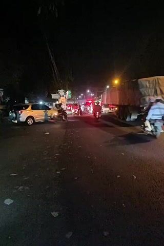 The video captures a nighttime scene on a road, likely in a city. The road is paved and appears to be busy with traffic. Several vehicles are visible, including a silver SUV on the left, a large truck with a covered cargo area on the right, and multiple motorcycles and smaller vehicles in the center. The vehicles are stopped or moving slowly, suggesting a traffic jam. Streetlights and vehicle headlights illuminate the scene, creating a somewhat hazy atmosphere. The surrounding environment is dark, with trees and buildings barely visible in the background. People are riding on the motorcycles, and some appear to be standing near the vehicles in the center. The overall impression is one of a typical evening commute or a busy intersection at night.