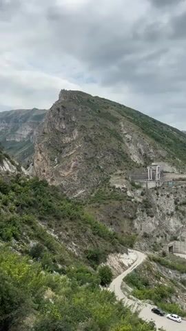 A drone shot pans from left to right over a mountainous, green landscape with a large body of turquoise water. A winding dirt road cuts through the scene. On the right side of the frame, a modern, green-roofed building with white walls sits on the edge of the water. Above it, a concrete structure is visible on the mountainside. In the distance, more rugged mountains are visible under a cloudy sky. Several cars are parked on the dirt road in the foreground.