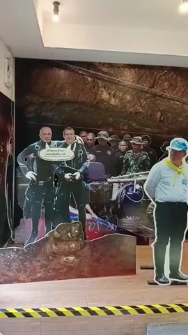 The video shows a static display of cardboard cutouts of people. The background appears to be a printed image of a cave or rock formation. In the foreground, there are several life-size cutouts. On the left are two cutouts of divers in full gear. Next to them is a cutout of a man in a light blue shirt and a blue hat with a yellow scarf. To his right are two cutouts of men in camouflage military uniforms. To the far right, there are partial cutouts of more figures. The cutouts are standing on black metal stands. There is a yellow and black striped border on the floor in front of the display.