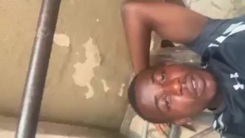 A man is visible in the video, lying on his back. He wears a black shirt and his hand is on his head. He is close to a wall with peeling paint. A metal pole is positioned beside him, and there is a tiled floor near him. The camera angle suggests it is low to the ground.