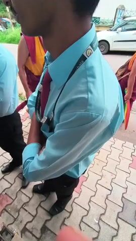 The video shows a street scene with several people. In the foreground, a person wearing a light blue shirt and a maroon tie is standing and looking to the left. Another person in similar attire is partially visible on the right. In the background, a Buddhist monk in maroon robes is walking away from the camera, carrying a bag and holding a multi-colored umbrella. Two white cars and a motorcycle are parked on the side of the road. The ground is paved with reddish-brown and light gray interlocking bricks. Vegetation is visible on the left side of the frame.