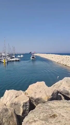 The video shows a clear blue sky over a body of water with several sailboats docked on the left side. A narrow channel, flanked by a rock barrier on the right, leads out to the open sea. A motorboat is moving away from the camera down this channel, leaving a wake behind it. The water is a deep blue, and the sunlight reflects off its surface. In the distance, a larger ship can be seen on the horizon. The foreground is dominated by large, rough-hewn rocks.