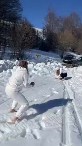Two women are in a snowy landscape on a sunny day. The woman on the left, with short brown hair, is wearing a white sweatsuit and tan-colored slip-on shoes. She is facing the other woman, holding something small in her hands, possibly a snowball. The woman on the right, with long dark hair and wearing a white hat and a black and white sweatsuit, is crouched down, gathering snow with her hands. In the background are bare trees, a small patch of green vegetation, and a couple of parked cars. The ground is covered in snow, with tire tracks visible. The sky is clear and blue.