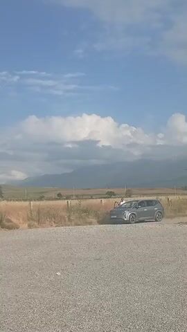 This media file captures a scene in Beş-Küŋgöy, Kyrgyzstan, on a bright, cloudy day. A modern, silver SUV is parked on a gravel surface next to a dry, grassy field. A person, wearing a white shirt, is standing near the open driver's side door of the SUV, seemingly engaged with the vehicle. In the background, a range of mountains is visible, partially obscured by dramatic, puffy white clouds against a vibrant blue sky. A lone utility pole stands in the middle ground, suggesting a rural or semi-rural setting. The dry vegetation hints at a warm season. The overall atmosphere appears calm and leisurely, with the activity centered around the parked vehicle and its occupant.