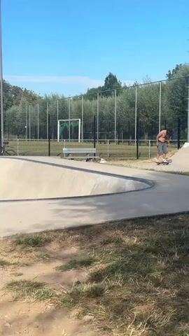 A person wearing gray shorts and no shirt stands on the edge of a concrete skatepark. In the background, trees and a fence are visible under a clear blue sky. Another person in a red shirt is further away, near a trash can.
