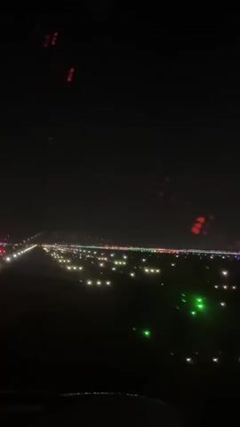 The video is taken from the perspective of an airplane window at night. The runway lights are visible, with white and green lights indicating the runway and red lights marking the edges. The lights stretch into the distance, disappearing into the darkness. The scene is dark, with only the runway lights providing illumination. There are some blurry red lights visible at the top of the frame, possibly from reflections on the window or lights from other aircraft. The video appears to be a short clip, possibly showing the plane during takeoff or landing.