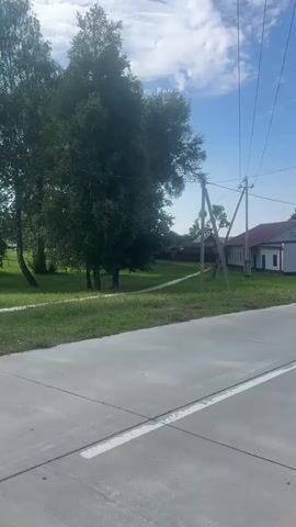 The video shows a paved road passing through a rural area. On the left side of the road, there is a white building with a red roof, surrounded by trees. Further down the road, a wooden pole with electrical wires is visible. On the right side of the road, a red wooden fence encloses a grassy area with a yellow house. The sky is blue with some scattered clouds. The video appears to be filmed from a moving vehicle.