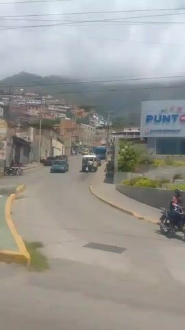 This video captures a daytime view of a street scene in La Guaira, Venezuela. The setting appears to be a moderately urban area with a mix of residential buildings, some appearing newer and well-maintained, while others look older and in disrepair. A prominent feature is a bright yellow building with a mural depicting a person's face, and text that is difficult to discern. Further down the street, another building, also painted yellow, has a more modern facade with multiple windows. A large billboard for "BUDA" is visible, featuring images of food, likely an advertisement for a restaurant or food establishment.

The street is paved and has yellow and green markings. Vehicles, including cars and a bus, are visible on the road, suggesting moderate traffic. A motorcycle is parked on the side of the street. In the background, a hillside with densely packed houses is visible, characteristic of many Venezuelan cities. The sky is overcast, indicating cloudy weather. There are utility poles with numerous wires crisscrossing overhead. The overall impression is a slice of everyday life in a Venezuelan town, with a blend of commercial activity and residential areas. The time of day appears to be midday or early afternoon due to the lighting conditions.